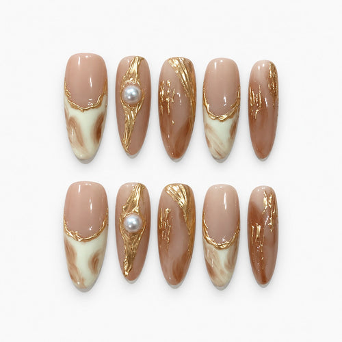 catalog: Long almond press-on nails in warm nude with cream and caramel marbling, raised gold foil lines, and a single cream pearl accent.