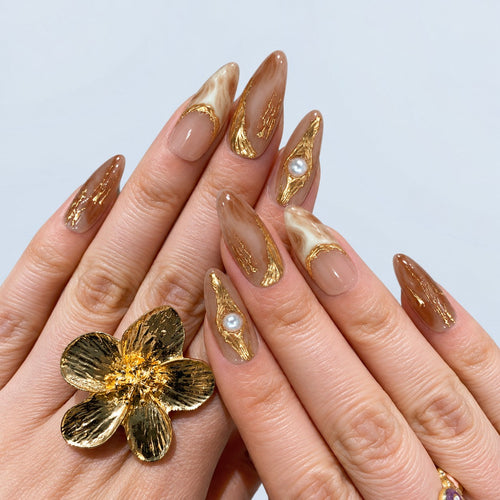 hand: Long almond press-on nails in warm nude with cream and caramel marbling, raised gold foil lines, and a single cream pearl accent.