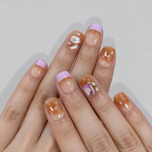 hand: Short square press-on nails in warm amber with lilac French tips, shell flecks and a pink rectangular gem.