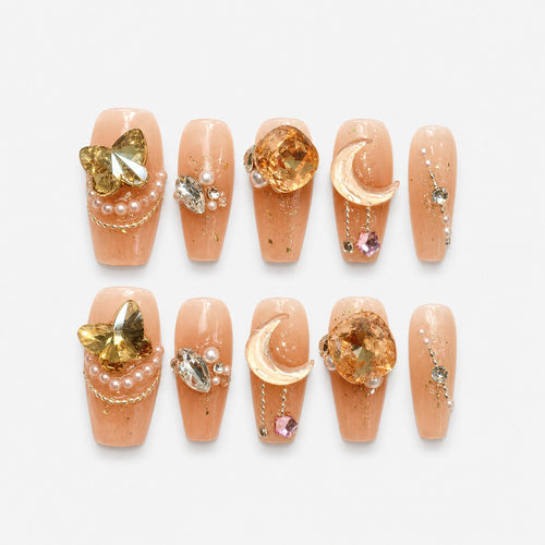 catalog: Medium nude press-on nails with gold flakes, an amber jewel, a pearl crescent moon on a chain, a gold butterfly and small pearls.