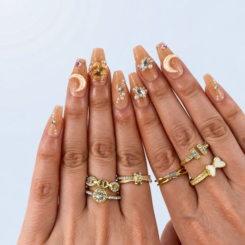 hand: Medium nude press-on nails with gold flakes, an amber jewel, a pearl crescent moon on a chain, a gold butterfly and small pearls.