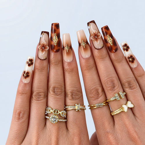 hand: Long square press-on nails in caramel and brown tortoiseshell with gold flower charms and painted cherries on a nude nail.