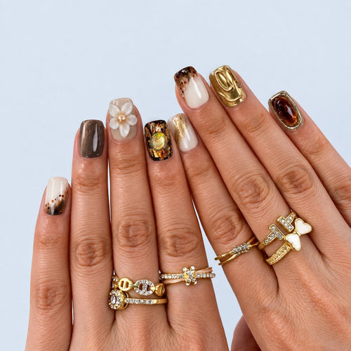 hand: Short square press-on nails in glossy brown cat-eye and milky white shades with gold foil streaks and one white sculpted flower with a pearl centre.