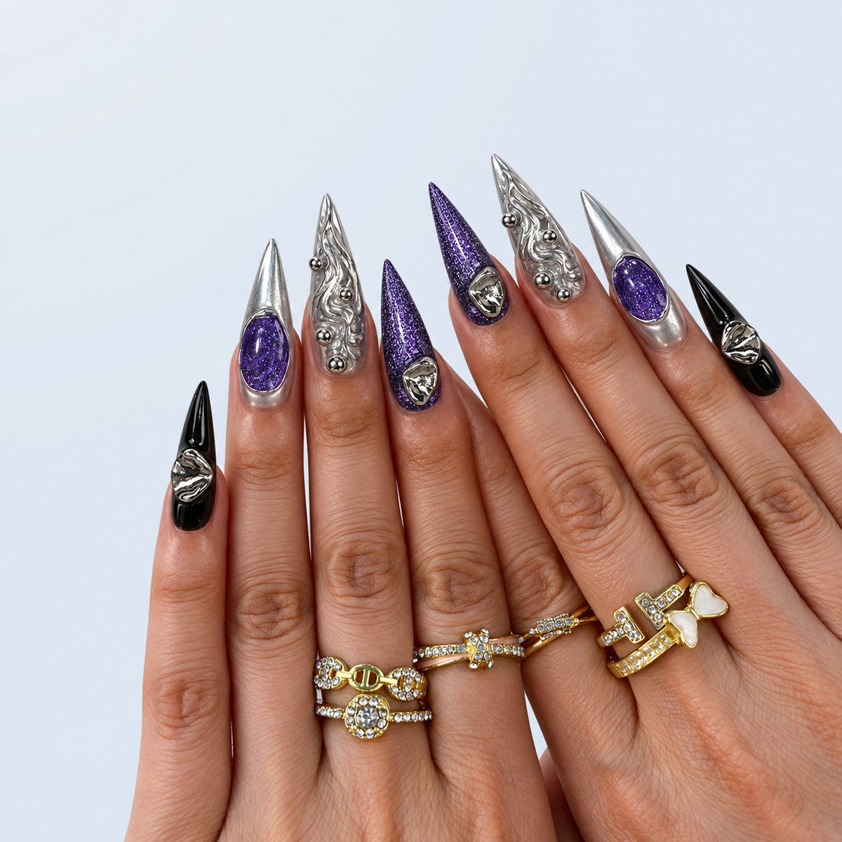 hand: Long stiletto press-on nails in mirror silver chrome with deep purple glitter stones, sculpted silver ripples with metal beads, and two glossy black accent nails.