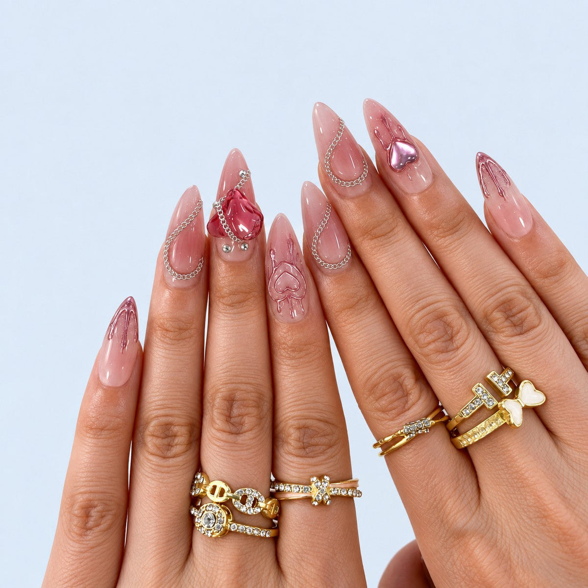 hand: Long almond press-on nails with a milky nude to dusty rose gradient, raised glass hearts, painted heart outlines, silver chains and small crystals.