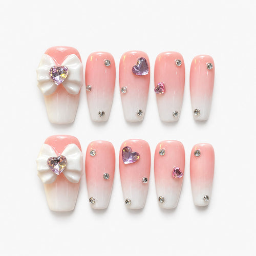catalog: Long coffin nails in a blush pink to white gradient with scattered clear crystals, lilac heart gems and a sculpted white bow.
