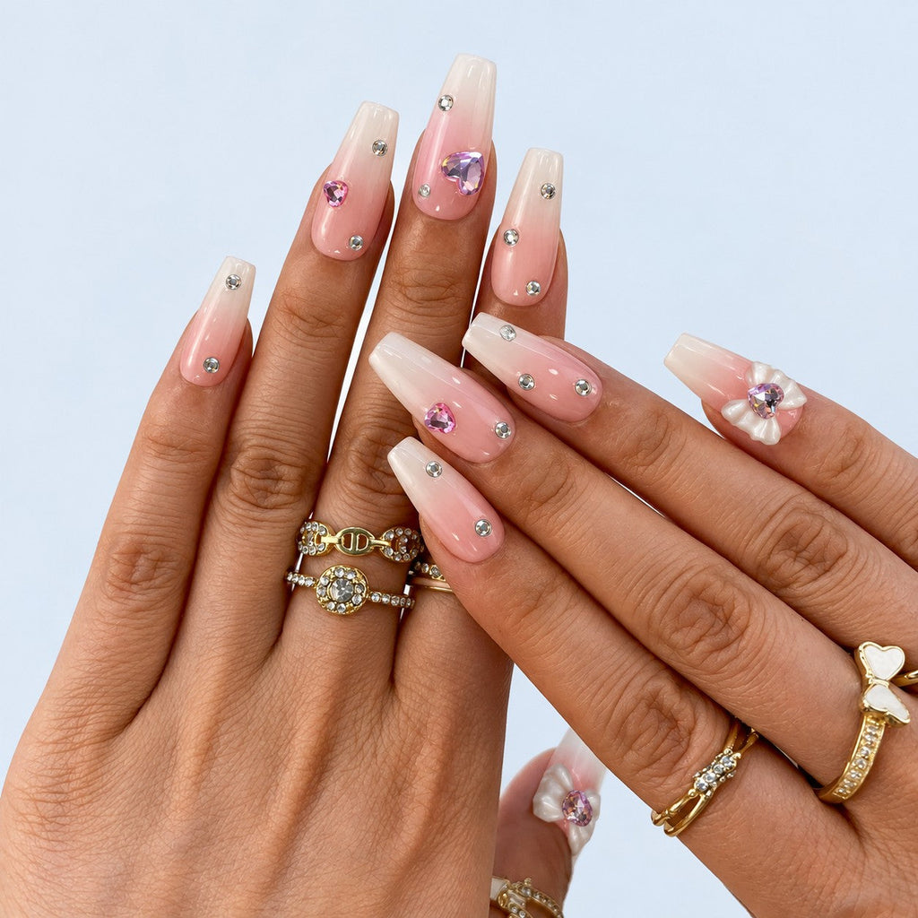 hand: Long coffin nails in a blush pink to white gradient with scattered clear crystals, lilac heart gems and a sculpted white bow.