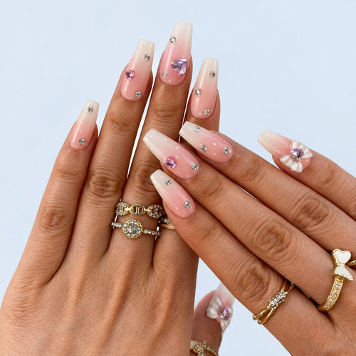 hand: Long coffin nails in a blush pink to white gradient with scattered clear crystals, lilac heart gems and a sculpted white bow.