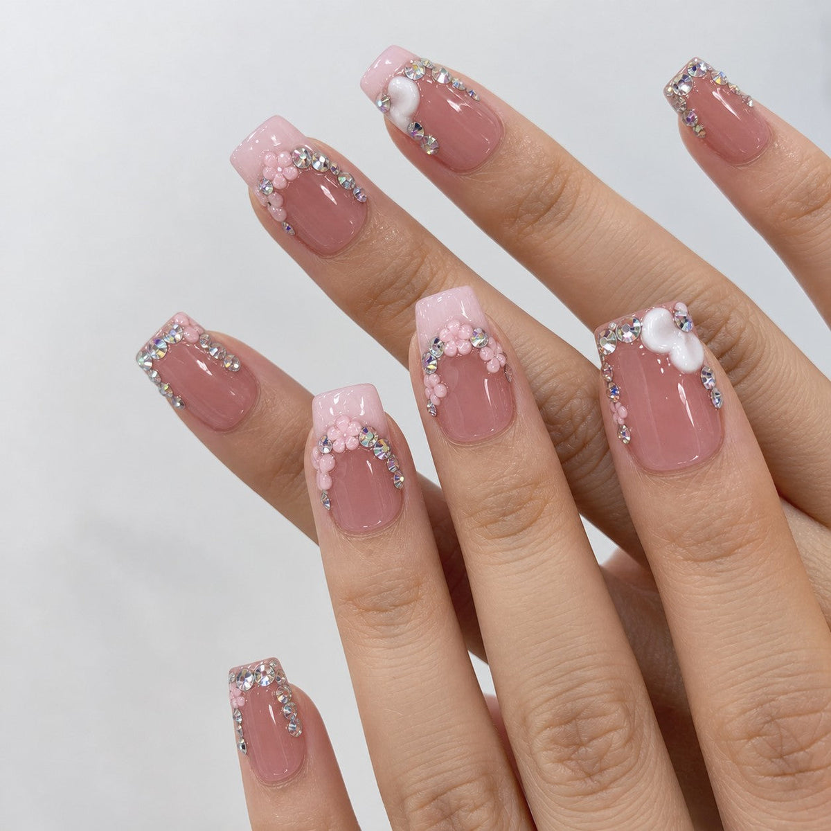 hand: Medium square press-on nails in dusty rose with a milky pink French tip, rhinestone clusters, sculpted pink flowers and white hearts.