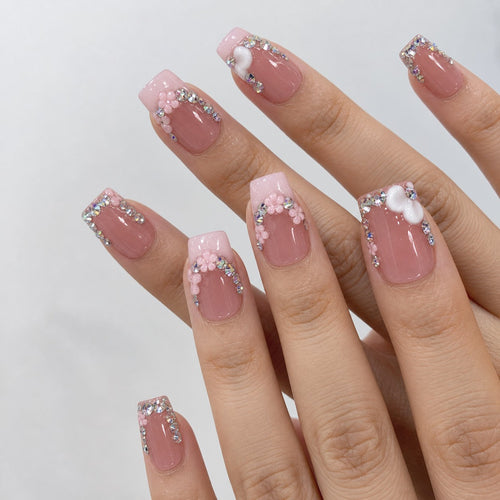 hand: Medium square press-on nails in dusty rose with a milky pink French tip, rhinestone clusters, sculpted pink flowers and white hearts.