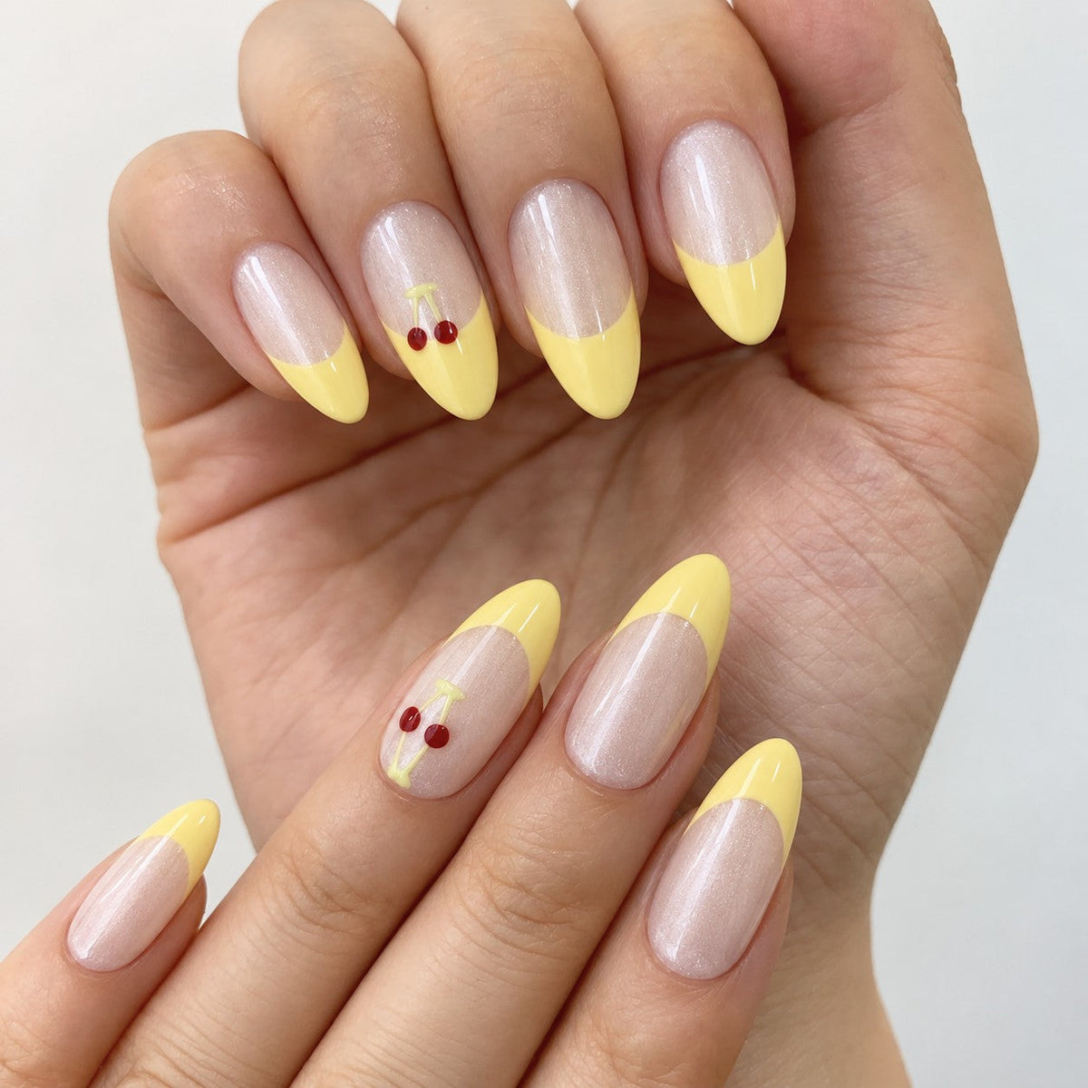 hand: Medium almond press-on nails with a pearly nude base, pastel yellow French tips and small red painted cherries on one accent nail.