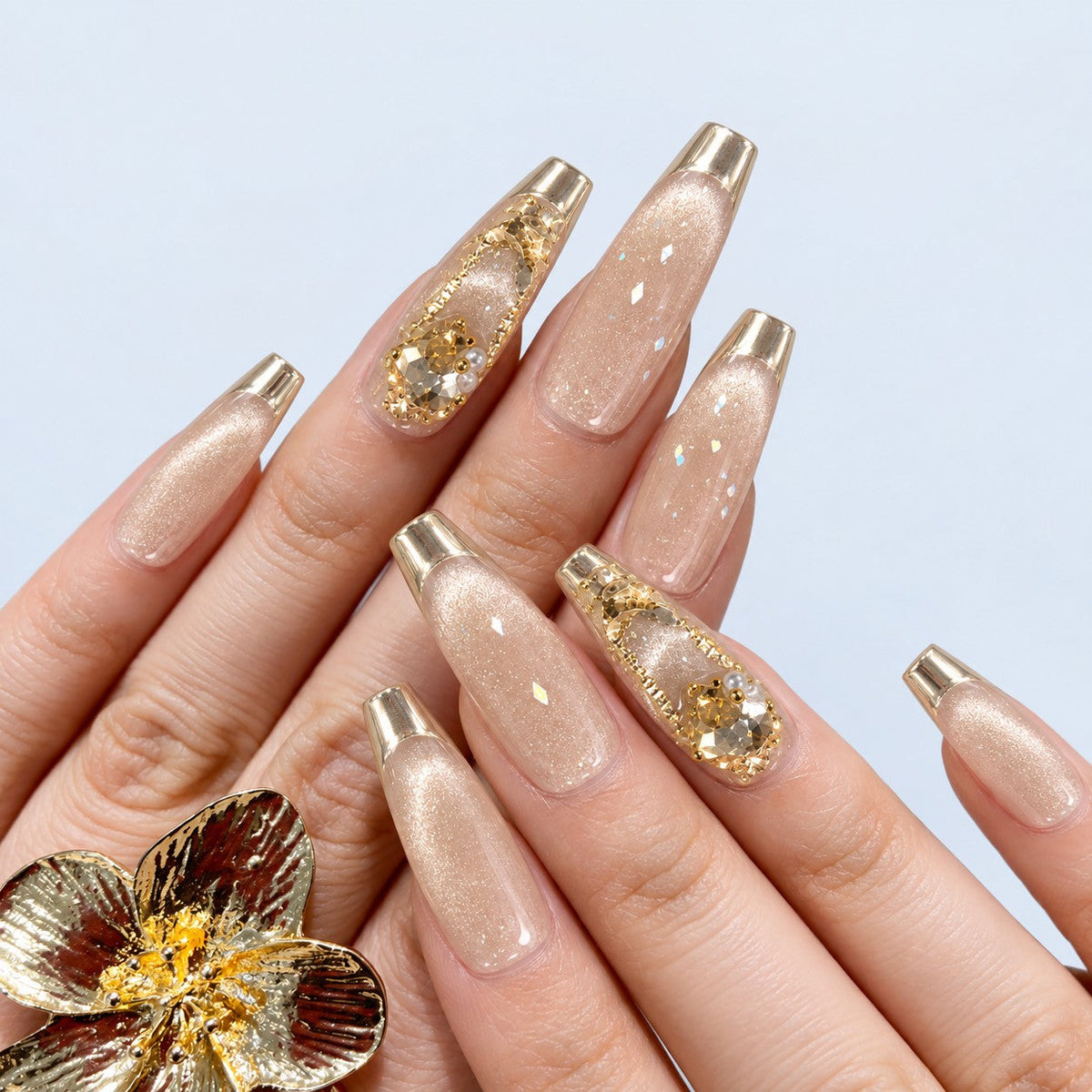 hand: Long coffin press-on nails in shimmering champagne with gold chrome French tips and raised gold gem accents.