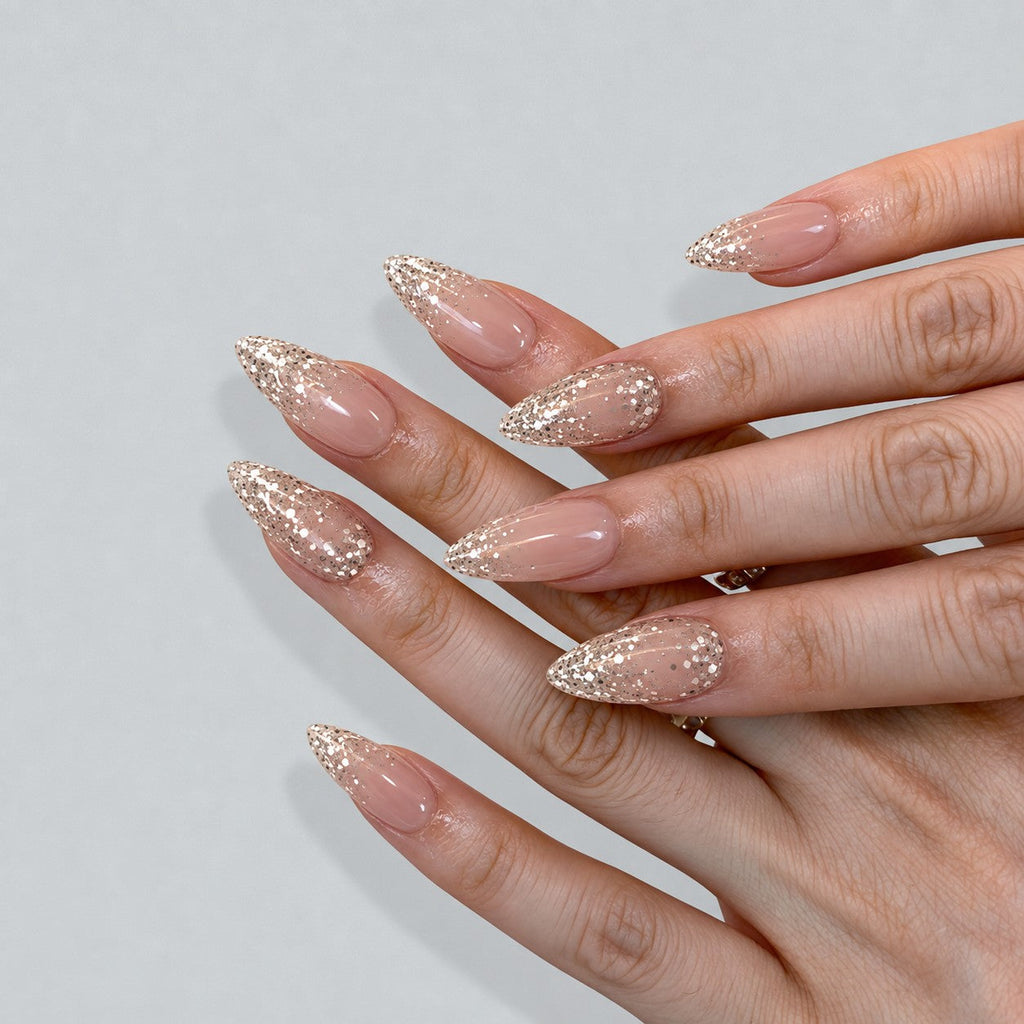 hand: Long almond press-on set in sheer nude with pearly shimmer and champagne gold glitter tips.