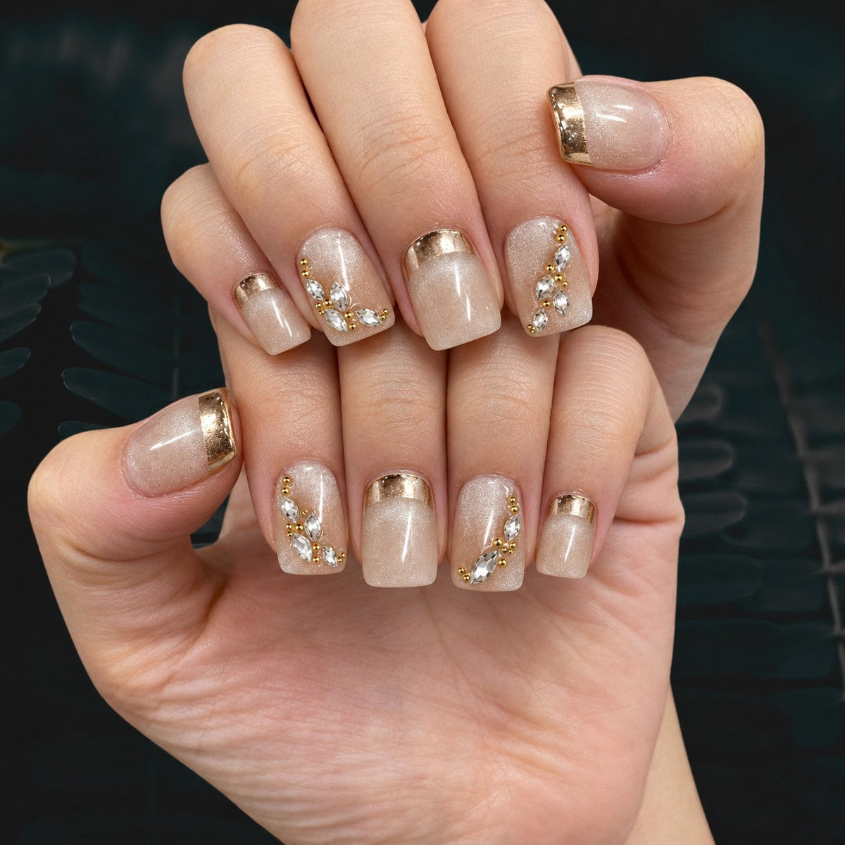 hand: Short square press-on nails in sheer nude with cat-eye shimmer, mirrored gold French tips and small clear crystal and gold bead accents.