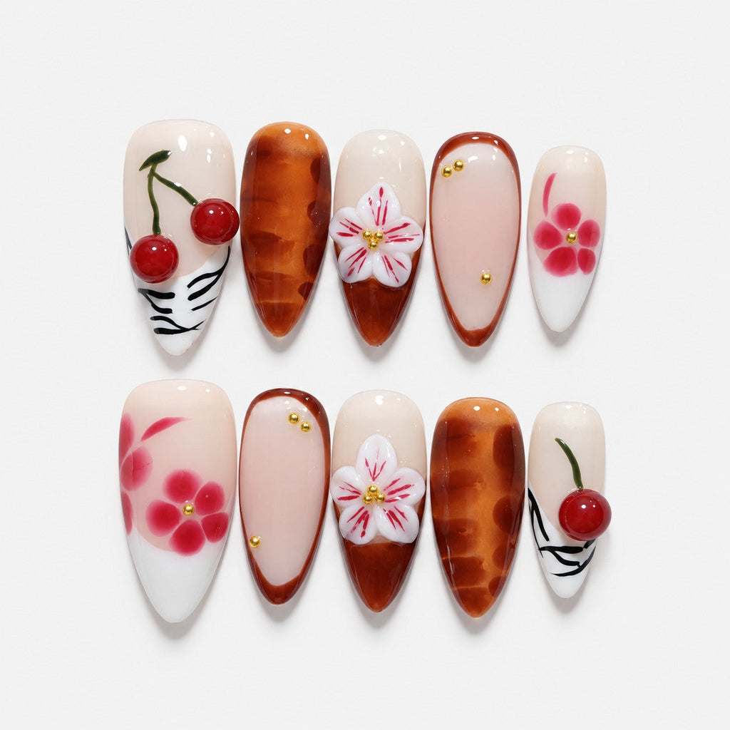 catalog: Long almond press-on nails in cream and amber brown with 3D red cherries, sculpted white lilies and painted pink blossoms.