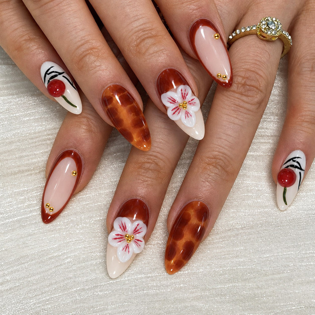 hand: Long almond press-on nails in cream and amber brown with 3D red cherries, sculpted white lilies and painted pink blossoms.