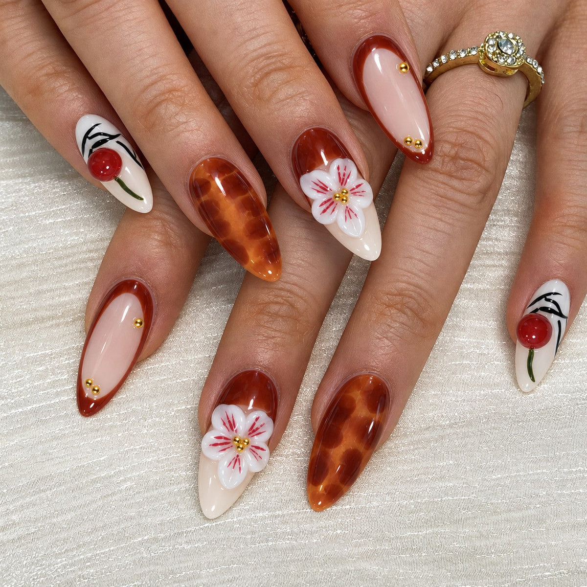 hand: Long almond press-on nails in cream and amber brown with 3D red cherries, sculpted white lilies and painted pink blossoms.