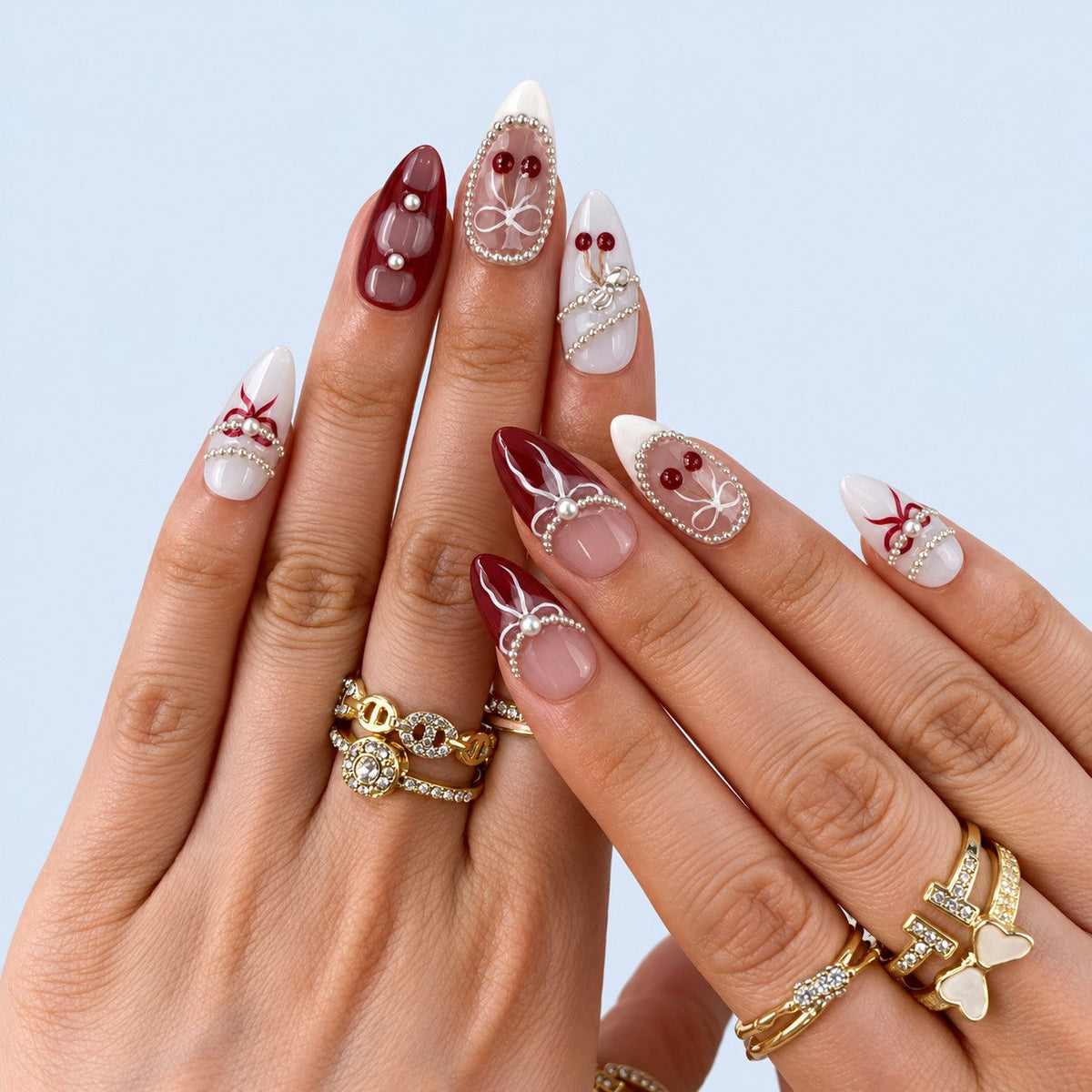hand: Medium almond press-on nails in burgundy, mauve and grey with painted white bows, silver bead strands and glossy red cherry charms.