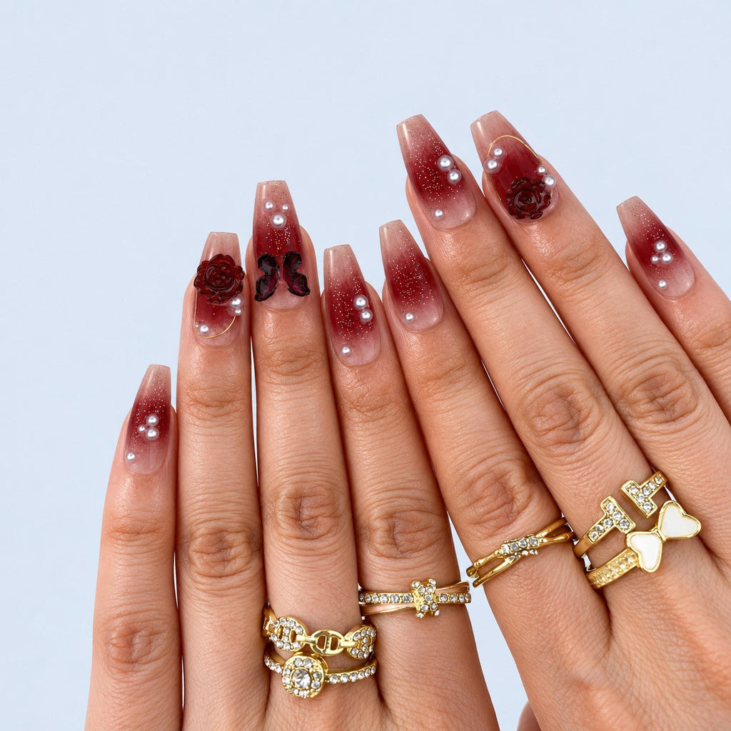 hand: Coffin shaped press-on nails with a wine red to nude gradient, sculpted dark red roses, a carved butterfly, grey pearls and fine gold lines.
