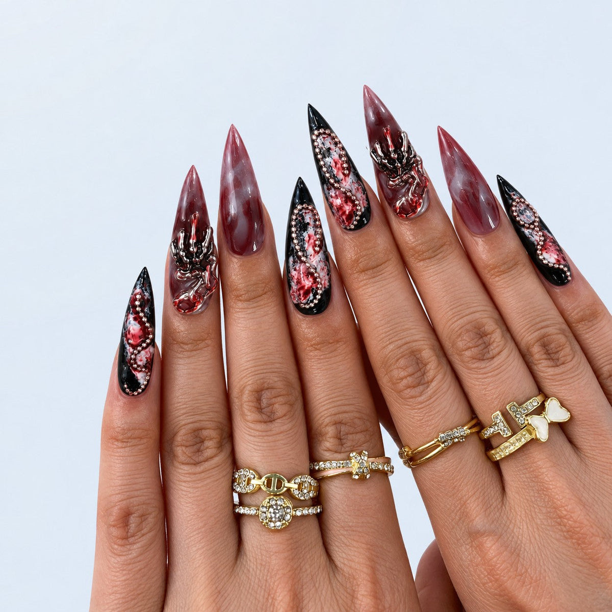 hand: Long stiletto press-on nails in blood red marble with sculpted roses, pearls, rose gold frames, silver skeleton hands and glossy black tips.
