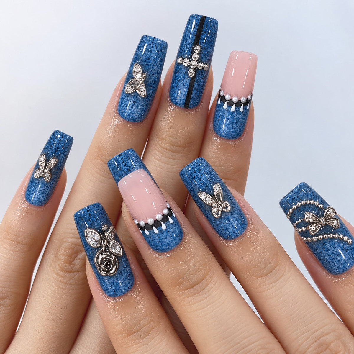 hand: Long square press-on nails in speckled denim blue with sheer pink accents, silver rose and bow charms, pearls and small crystals.