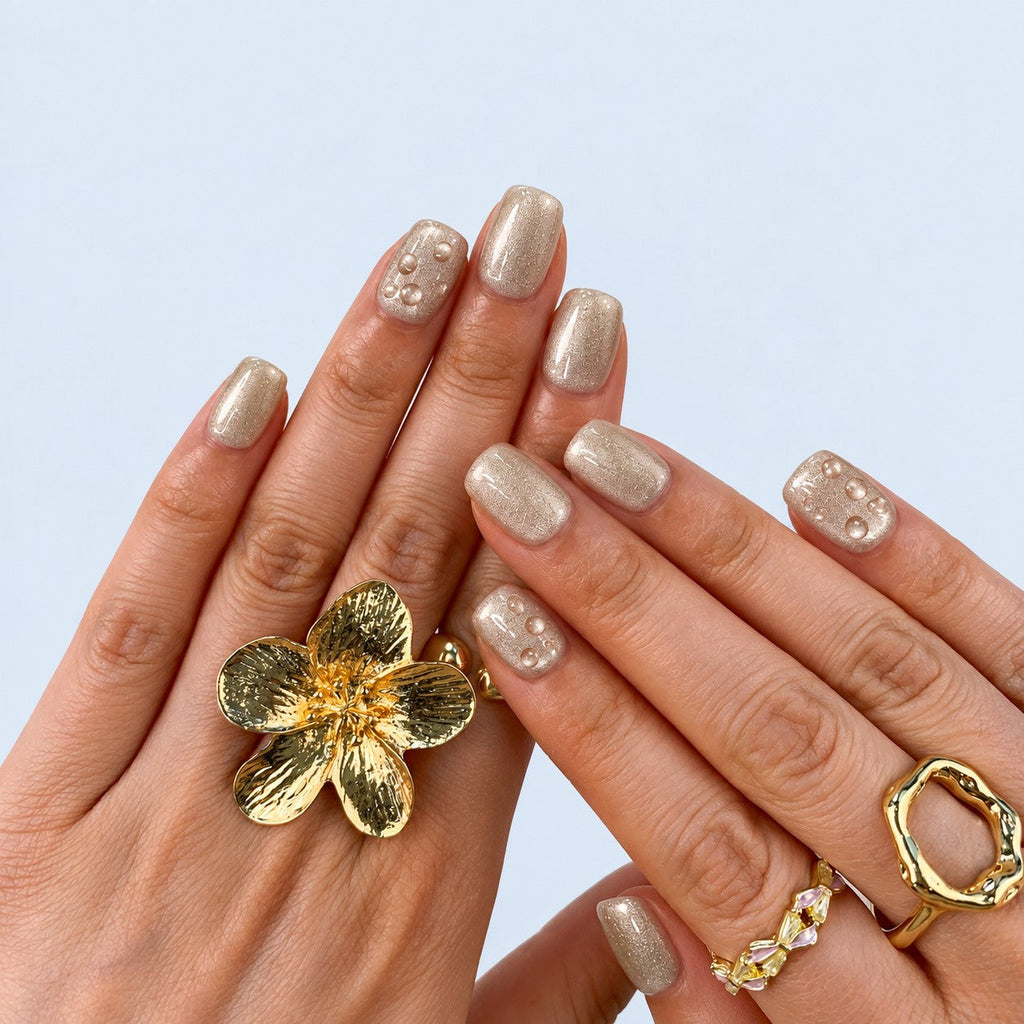 hand: Short square press-on nails in frosted champagne pearl with small raised clear droplets.
