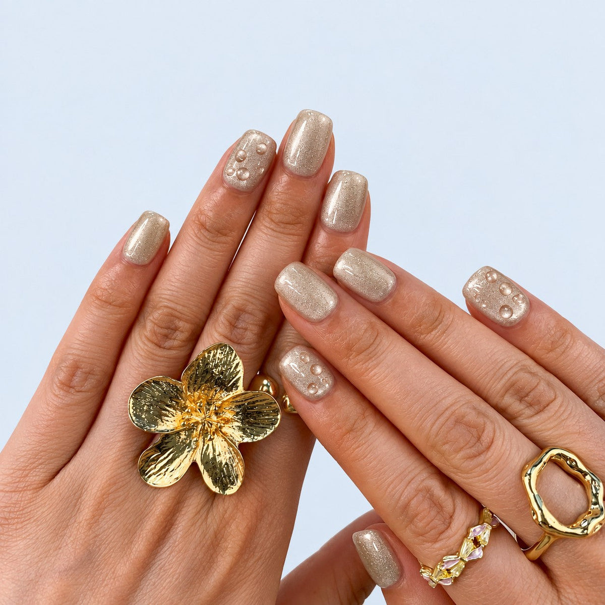 hand: Short square press-on nails in frosted champagne pearl with small raised clear droplets.