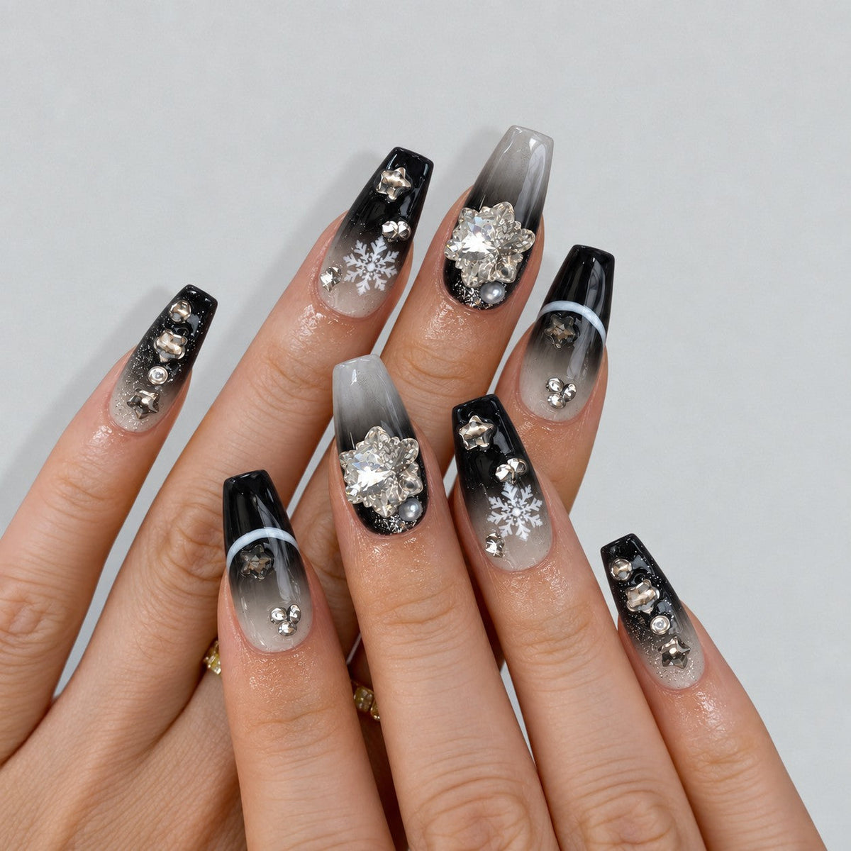 hand: Coffin shaped press-on nails with black and grey gradient, silver glitter, crystal snowflake charms and mirrored star studs.