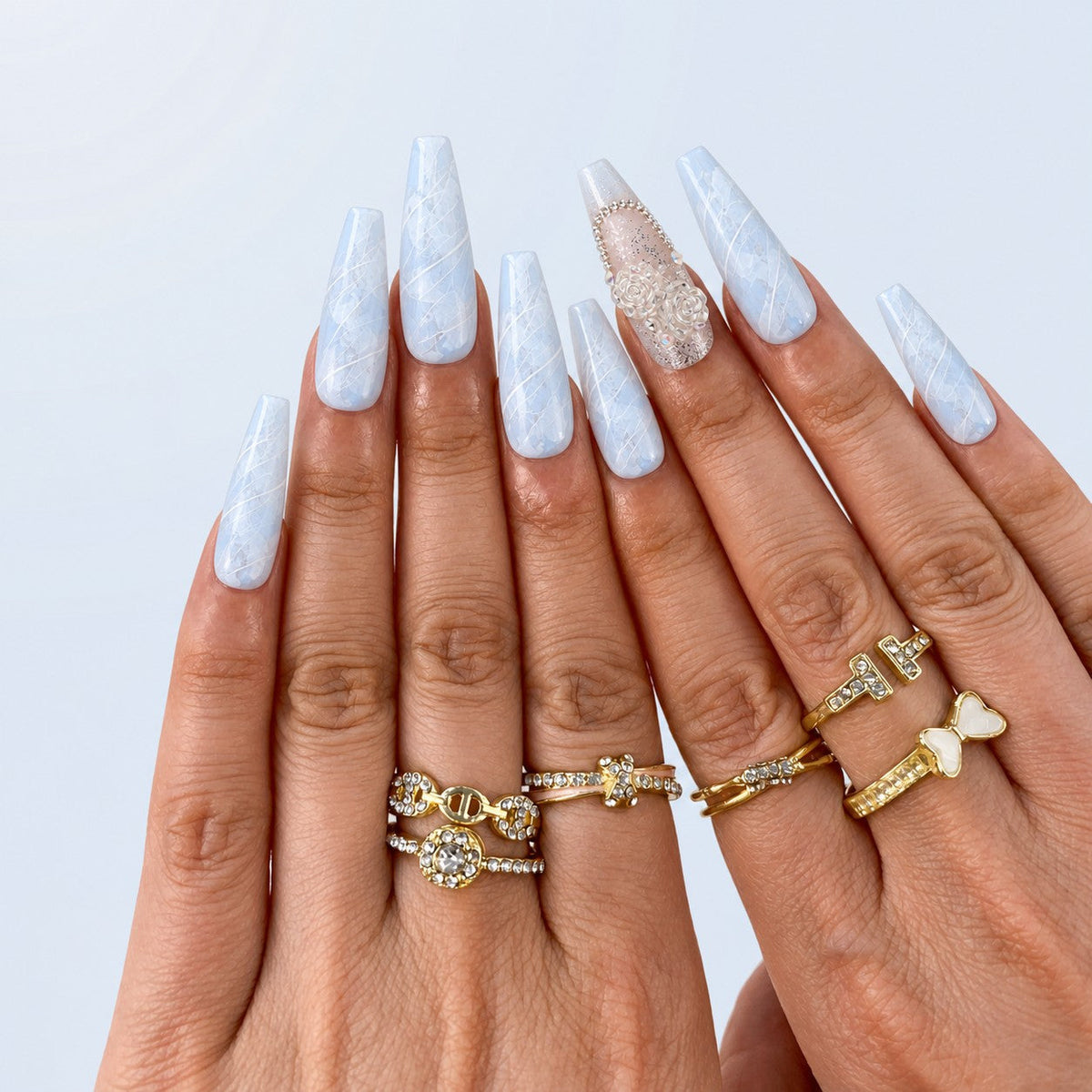 hand: Long coffin press-on nails in blue and white marble with silver line detail and two accent nails holding sculpted white roses and crystals.
