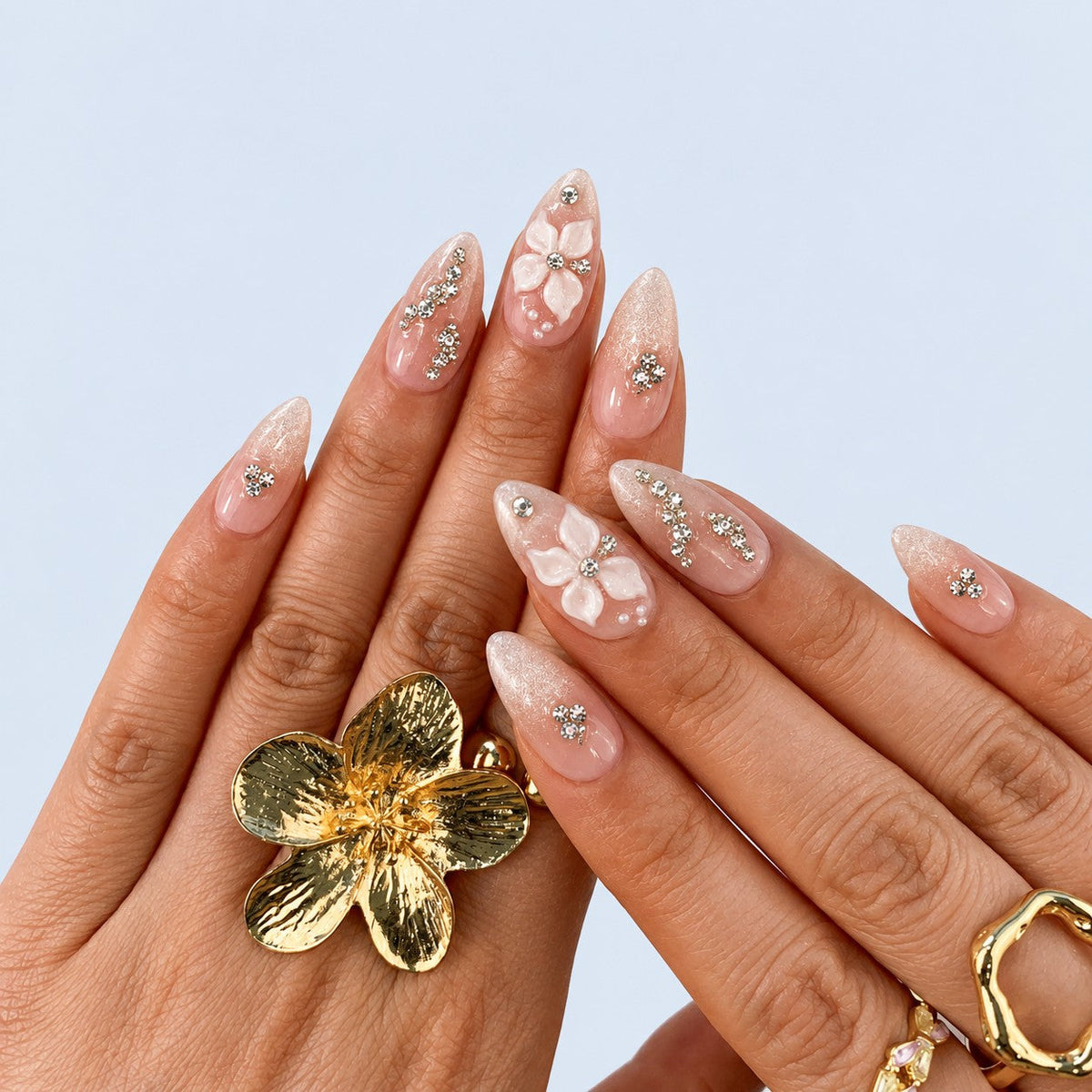 hand: Almond medium press-on nails in milky nude with pearl shimmer, sculpted white 3D flowers and clear rhinestone accents.