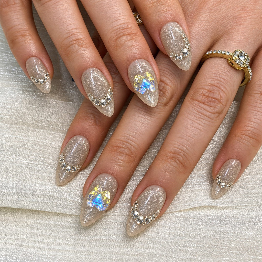 hand: Medium almond press-on nails in icy greige cat-eye shimmer with clear crystal curves, a star gem and a large iridescent heart gem.