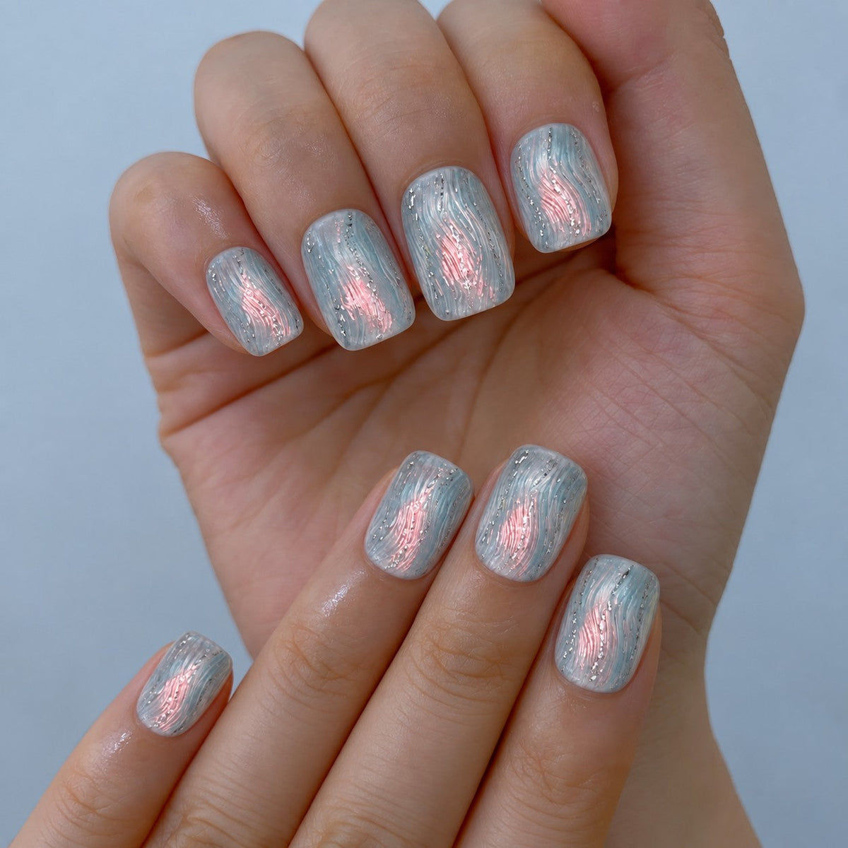 hand: Short square press-on nails in frosted ice blue with textured ripples, a pearly pink shimmer through the centre and silver glitter lines.