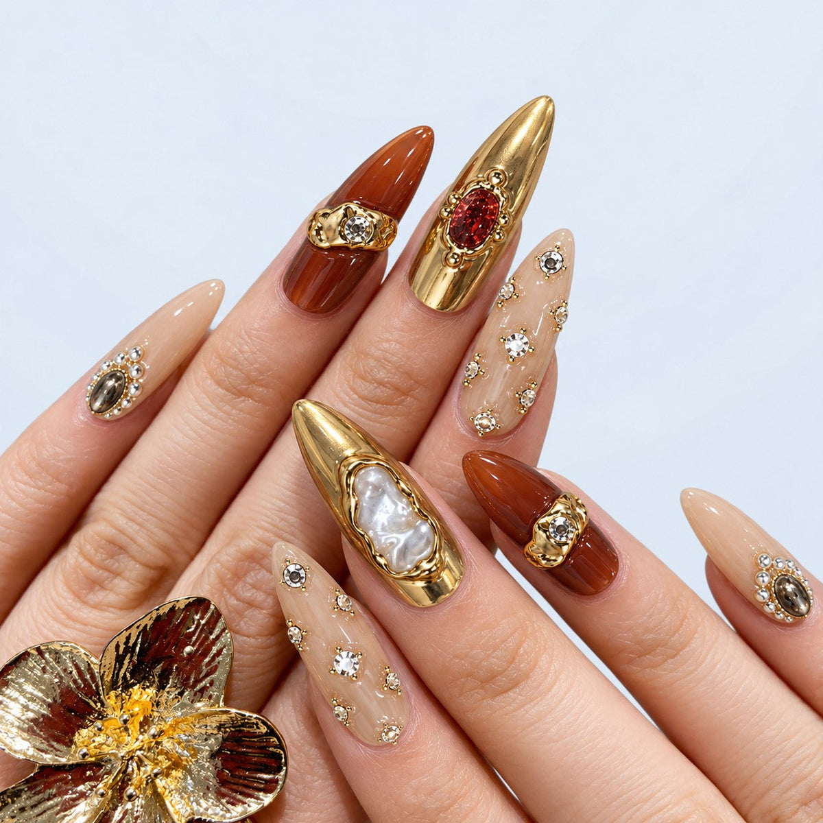 hand: Long almond press-on nails in gold chrome and amber brown with baroque pearls, crystals and raised gold sculpting.