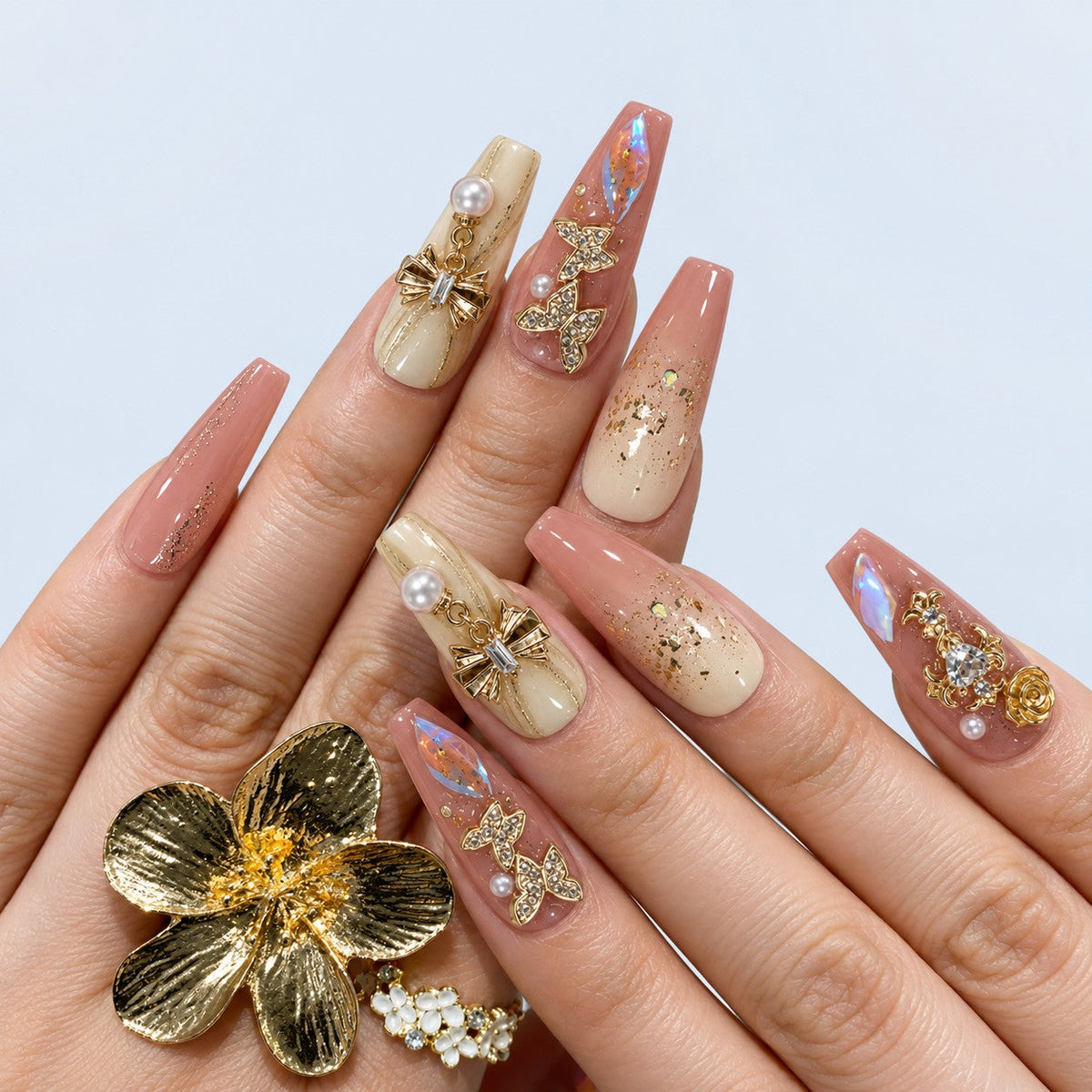 hand: Long coffin press-on nails in pink and ivory with gold leaf, rhinestone butterflies, a gold bow with a pearl drop and a crystal heart charm.