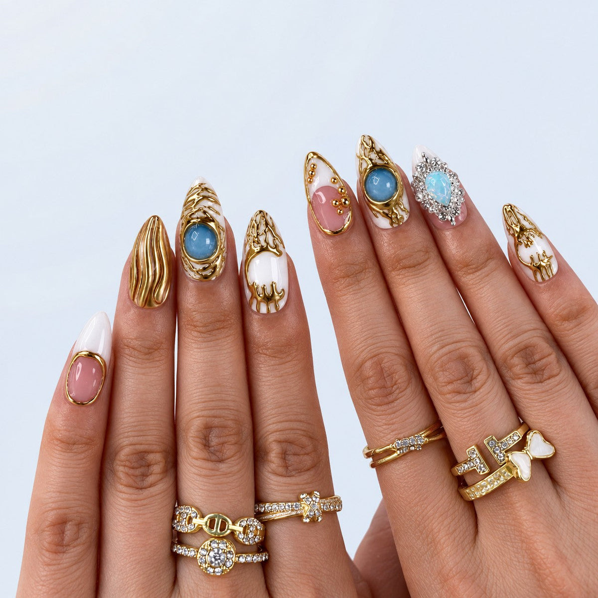 hand: Medium almond press-on nails in white and pink with raised molten gold work, blue cabochon stones, gold beads and a crystal set opal charm.
