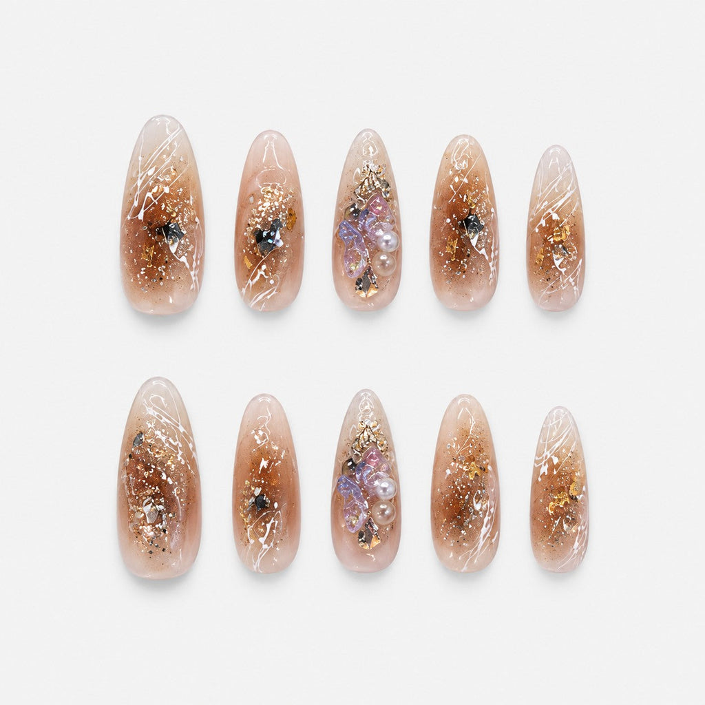 catalog: Long almond nails in sheer champagne with gold foil flakes, white line art, glitter and a lilac crystal and pearl cluster.