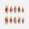catalog: Long almond nails in sheer champagne with gold foil flakes, white line art, glitter and a lilac crystal and pearl cluster.