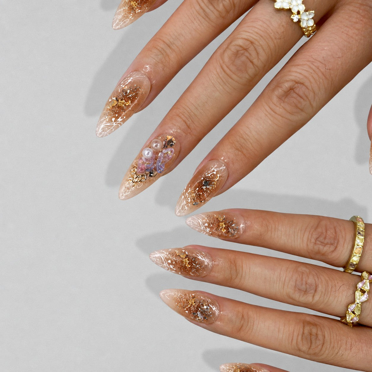 hand: Long almond nails in sheer champagne with gold foil flakes, white line art, glitter and a lilac crystal and pearl cluster.