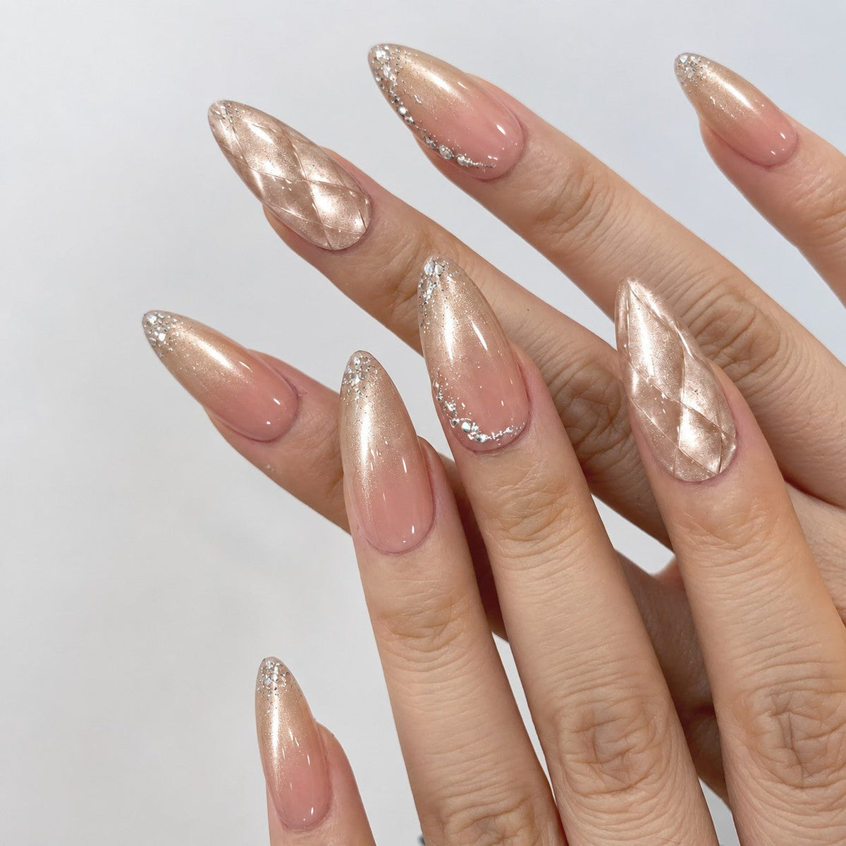 hand: Long almond press-on nails in nude pink fading to mirror champagne gold, one with a quilted diamond texture, plus glitter and small crystals.