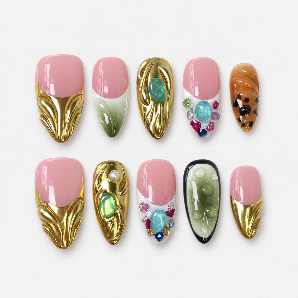 catalog: Medium almond press-on nails with soft pink beds, sculpted mirror gold French tips, green jelly stones, a painted leaf and clustered coloured gems.