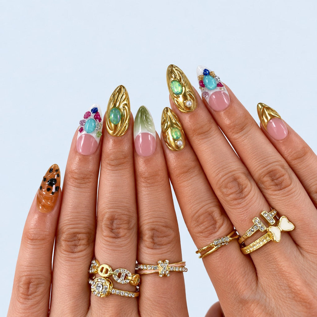 hand: Medium almond press-on nails with soft pink beds, sculpted mirror gold French tips, green jelly stones, a painted leaf and clustered coloured gems.