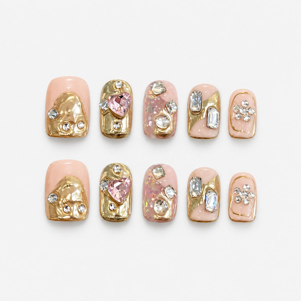 catalog: Short square press-on nails in peach nude with sculpted gold foil shapes and clusters of clear and pink crystals.