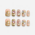 catalog: Short square press-on nails in peach nude with sculpted gold foil shapes and clusters of clear and pink crystals.