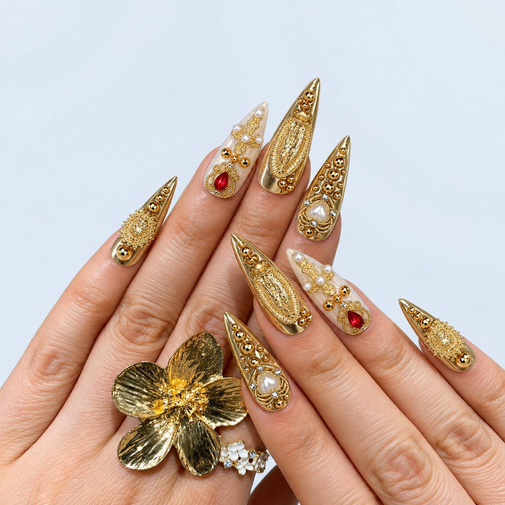 hand: Long stiletto press-on nails in mirror gold and pearl white with a crown charm, pearl drop, red teardrop crystal and ornate gold filigree.