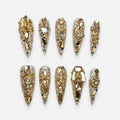 catalog: Long stiletto press-on nails covered in champagne glitter with gold baroque scrollwork, clear rhinestones and mirrored gold shards.