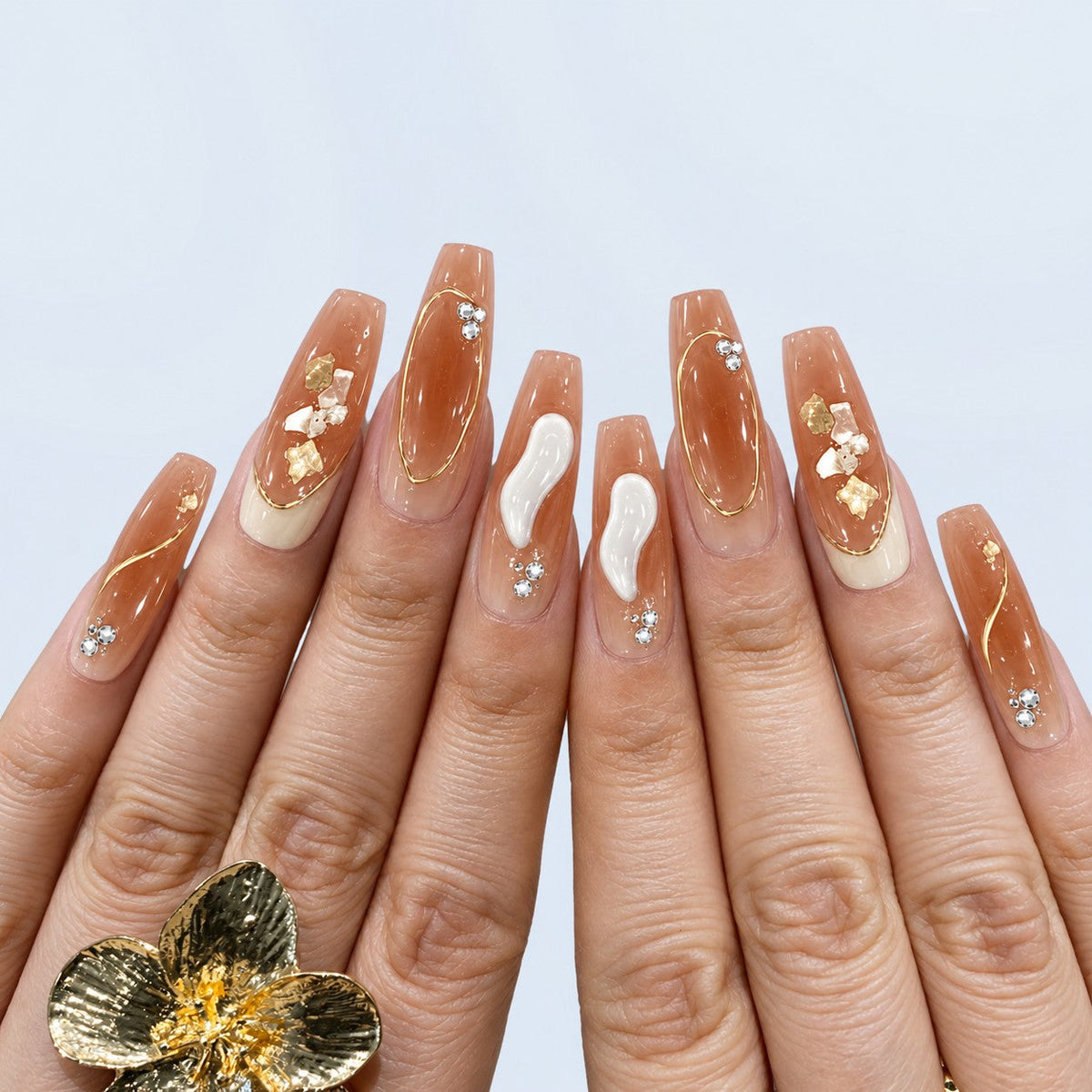 hand: Long coffin nails in sheer nude with gold foil outlines, small white flowers, cream tips and tiny crystals.
