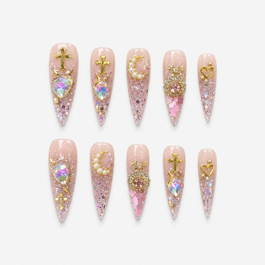 catalog: Long stiletto press-on nails in clear pink glitter with gold cross charms, a pearl crescent moon and large iridescent heart-shaped stones.