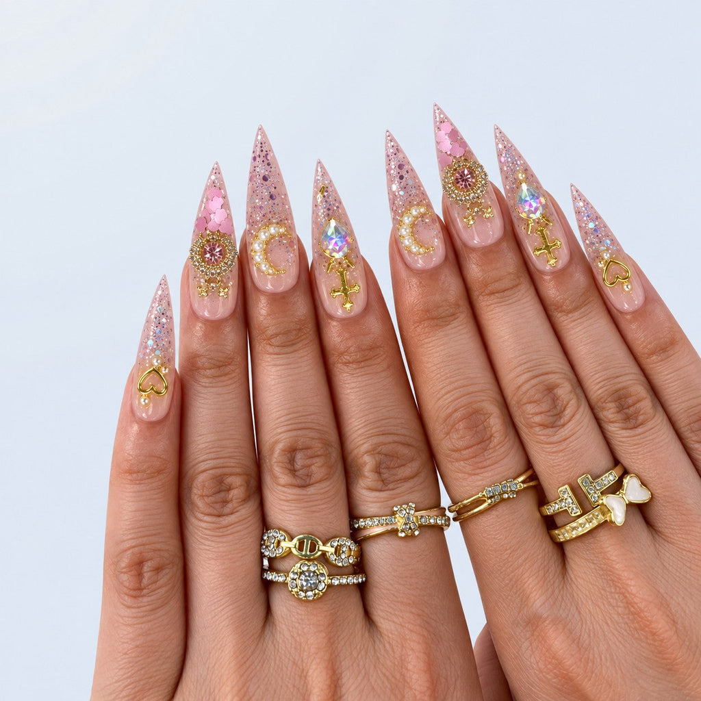 hand: Long stiletto press-on nails in clear pink glitter with gold cross charms, a pearl crescent moon and large iridescent heart-shaped stones.