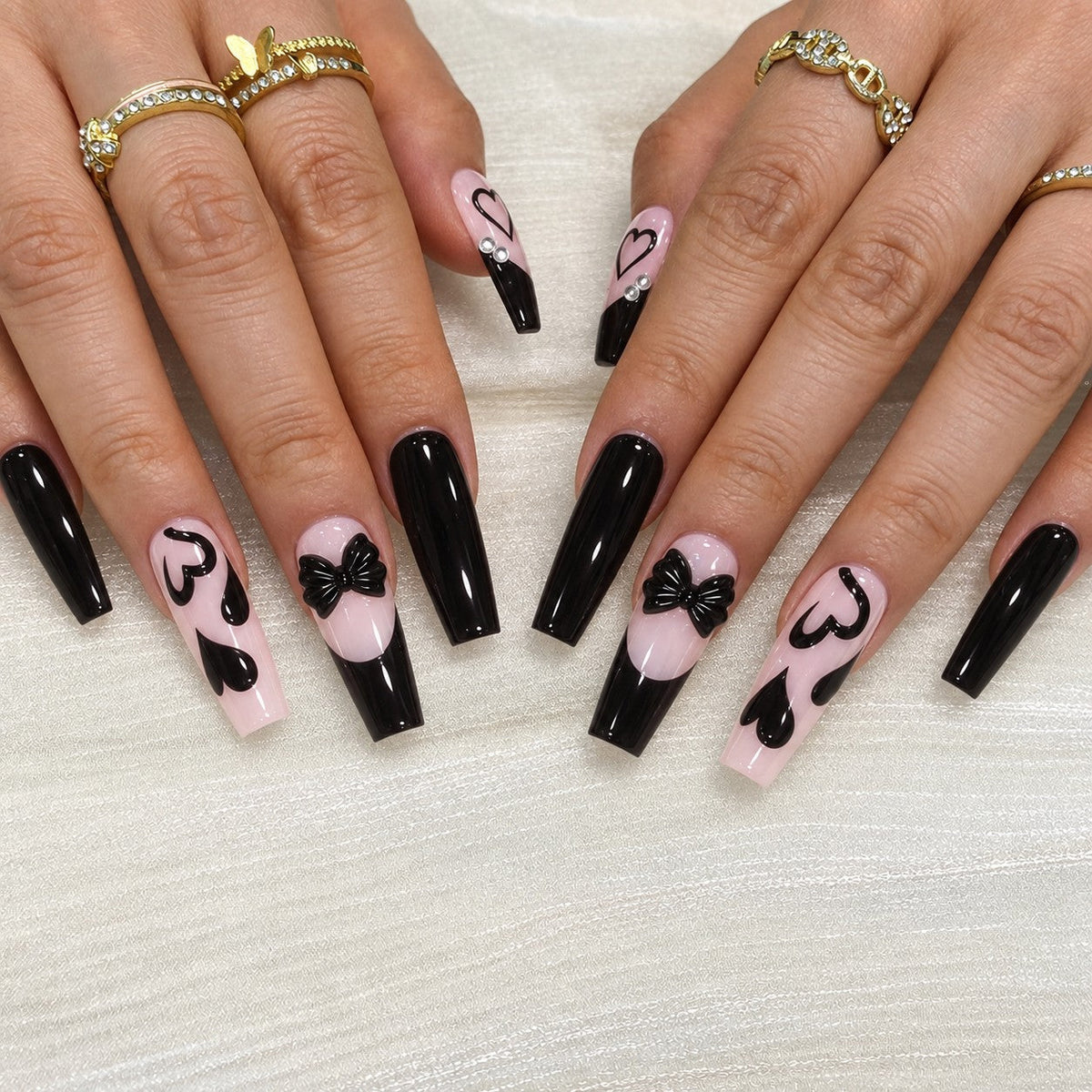 hand: Long coffin press-on nails in near black over soft pink with slanted French tips, painted hearts and a sculpted black bow.