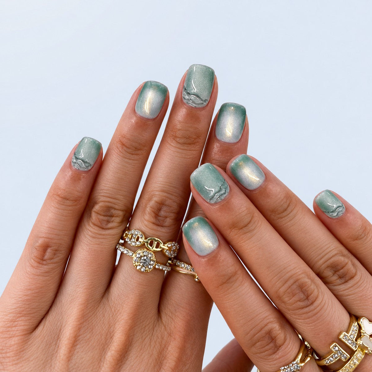 hand: Short square press-on nails in sage green with a pearly cat-eye shimmer and raised clear brushstroke detail.